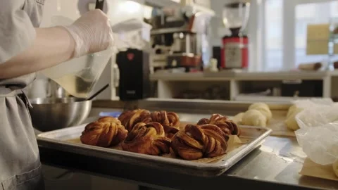 The baker uses a brush to apply syrup to freshly baked, hot cinnamon rolls. Stock Footage 267017288