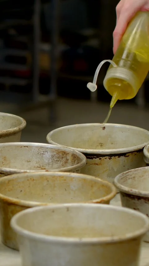 Bakery worker pouring oil into worn round tins on countertop with shallow Видео 344574304