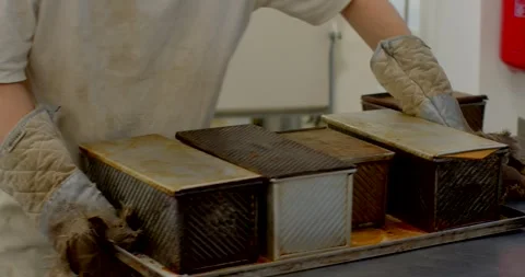 Bakery worker transferring hot loaf pans with quilted oven mitts in commercial Stock Footage 344582804