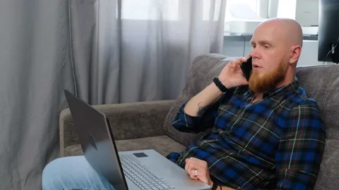 Bald guy with a laptop on his lap talking on the phone. Serious face Stock Footage 139191206