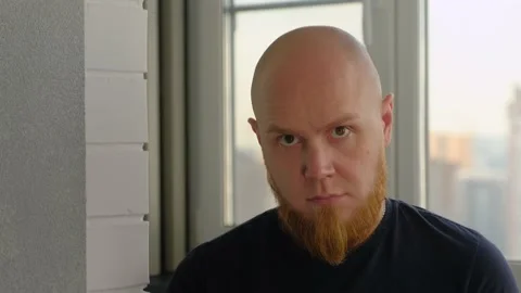 A bald guy sits by the window, looks at his feet, and then at the camera. Stock Footage 142580645