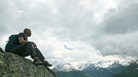 Bald man with a backpack sits on a rocky ledge, gazing at the expansive snow Stock Footage 282814449