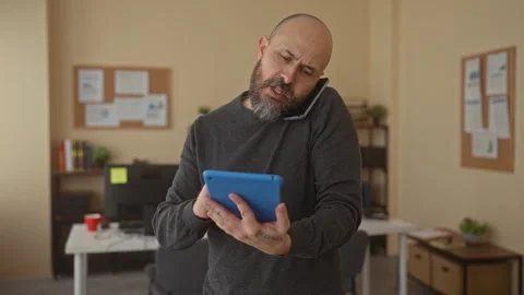 Bald man with beard multitasks in office holding tablet while on phone, sur.. Video stock 310989318