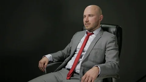 Bald man with a beard in a suit sits in a black leather chair looking to the Stock Footage