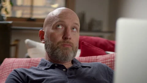Bald man with a beard thinks for inspiration while working on a laptop at home Video stock 133286456