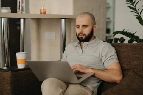 A bald man with a beard works remotely on his laptop sitting on a sofa at hom Stock Photos
