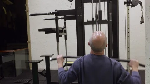 Bald man doing lat pulldown on cable machine with arms extended in home gym Stock Footage 324755557
