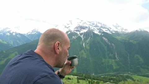 Bald man drinking from a metal flask while admiring a stunning mountain 스톡 동영상 282230765