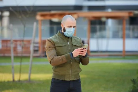 A bald man in a face mask checking his smartphone on a cozy street. Stock Photos