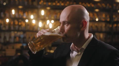 A bald man in a jacket is sitting at a table and drinking beer in a pub. The man Video stock 261092229