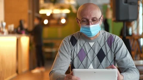 Bald man in mask sits at the table in restaurant and works on tablet computer Stock Footage 143425121