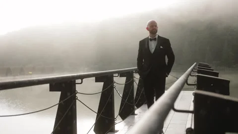 Bald man in a sharp black tuxedo stands confidently on bridge, with one hand in Stock Footage 284343389