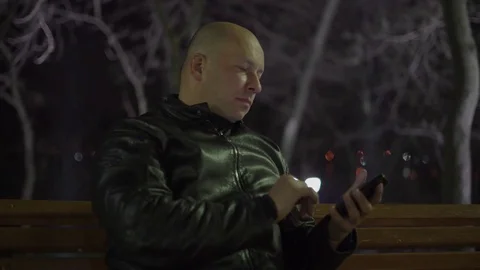 A Bald Man Sits on a Park Bench Doing Operations With His Mobile Phone in the Li Stock Footage 74949110