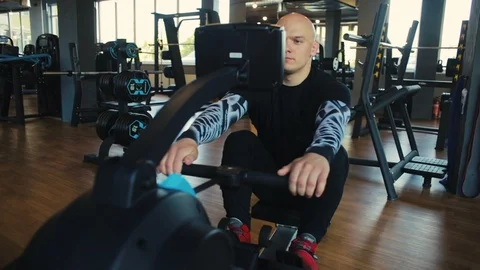 Bald man sits on rowing machine and pulls oar actively Stock Footage 110575068