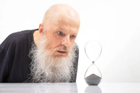 Bald man with a white beard closely examines an hourglass, symbolizing the .. Stock Photos