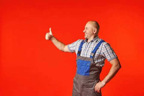 Bald man in work clothes on a red background shows the ok gesture to the side Stock Photos