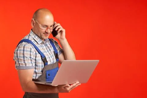 Bald man in work clothes talks on the phone and holds a laptop Foto stock