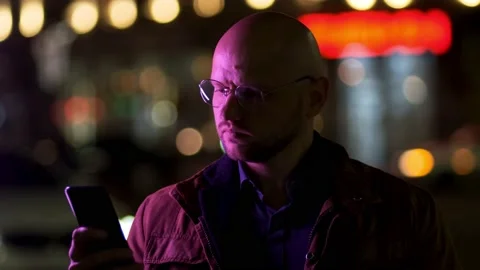 A bald, solid guy in a brown jacket uses a mobile phone on the street at night Stock Footage 195911484