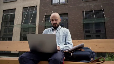 Bald young man freelancer with a beard sits on a wooden bench in urban space Stock Footage 134211101