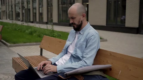 Bald young man freelancer with a beard sits on a wooden bench in urban space Stock Footage 134211176