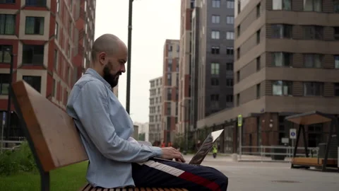 Bald young man freelancer with a beard sits on a wooden bench in urban space Stock Footage 134211289