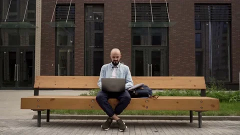 Bald young man freelancer with a beard sits on a wooden bench in urban space Stock Footage 135108015