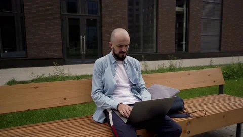 Bald young man freelancer with a beard sits on a wooden bench in urban space Stock Footage 135108057