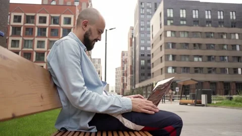 Bald young man freelancer with a beard sits on a wooden bench in urban space Video stock 135108070