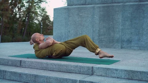 Bald Young Man Makes Complex Yoga Exercises in the City Park Lying on the Green Stock Footage 85330548