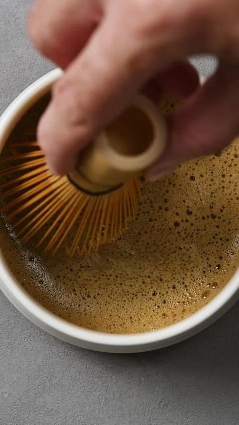 Bamboo whisk moves slowly across foamy hojicha surface in ceramic bowl, showing Stock Footage 343495341