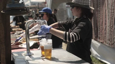 Bar girl in hat pours cold beers into plastic cup at sunny festival event Stock Footage 193370499