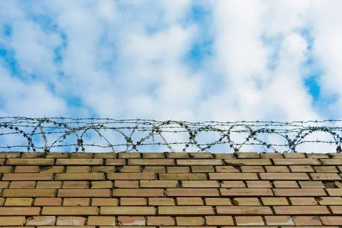 Barbed wire on a brick wall against a blue sky with clouds. Stock Photos