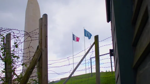 Barbed wire, two flags, and a tall monument set against a grassy hill Видео 299064668