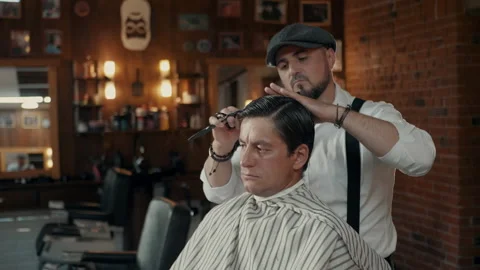 Barber with a beard and a cap cuts the client with scissors in a barbershop. Stock Footage 132284045