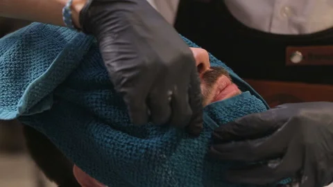 Barber covers the face of a man with a beard with a hot towel. Photo in vintage Stock Footage 93711689