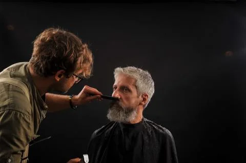 Barber cuts a beard to a client to an elderly gray-haired man Stock Photos