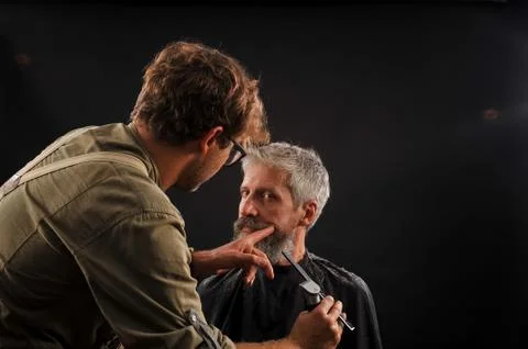 Barber cuts a beard to a client to an elderly gray-haired man Stock Photos
