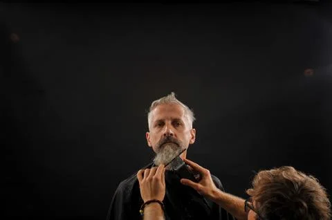 Barber cuts a beard to a client to an elderly gray-haired man Stock Photos