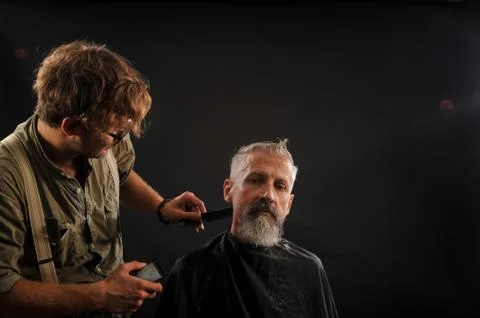 Barber cuts a beard to a client to an elderly gray-haired man Stock Photos