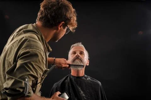 Barber cuts a beard to a client to an elderly gray-haired man Stock Photos