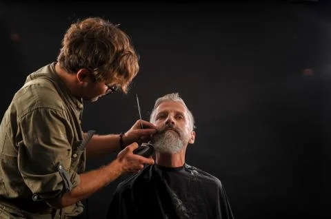 Barber cuts a beard to a client to an elderly gray-haired man Foto stock