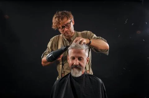 Barber cuts a beard to a client to an elderly gray-haired man Stock Photos