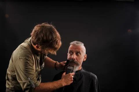 Barber cuts a beard to a client to an elderly gray-haired man Stock Photos