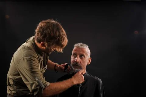 Barber cuts a beard to a client to an elderly gray-haired man Stock Photos