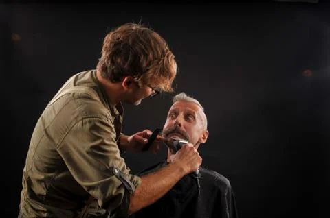 Barber cuts a beard to a client to an elderly gray-haired man Stock Photos