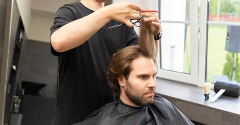 A barber uses scissors and a comb to give a haircut to a male client with lon Fotos Stock