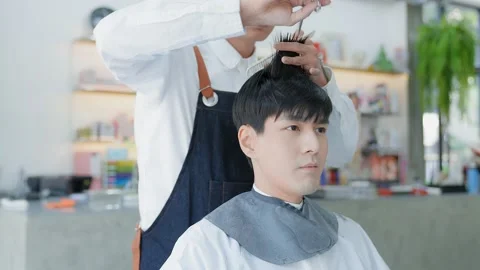A barber using scissors to cut an Asian male client's hair. Professional hair Stock Footage 153954364