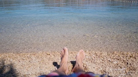 Bare feet of a man laying on a beach. Transparent sea waves wash stony shore 스톡 동영상 125640935