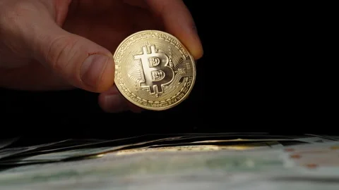Bare Hand Holding a Shiny Bitcoin Coin Over a Stack of US Dollar Bills Against a Stock Footage 296564878