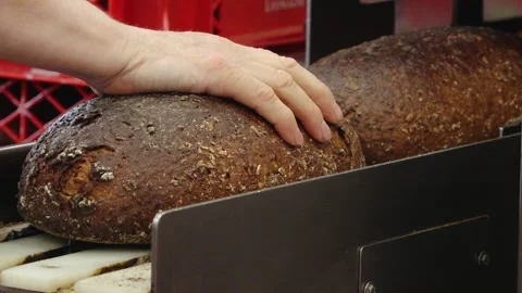 A bare hand steadies a large crusty loaf of dark bread on bakery slicing Stock Footage 335299014